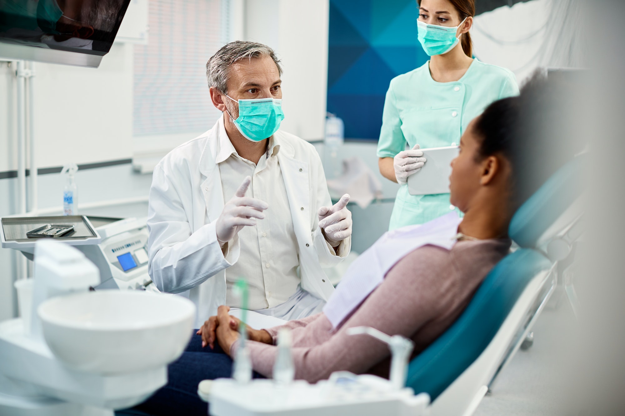 Mature dentist with face mask talking to black female patient at dentist's office.