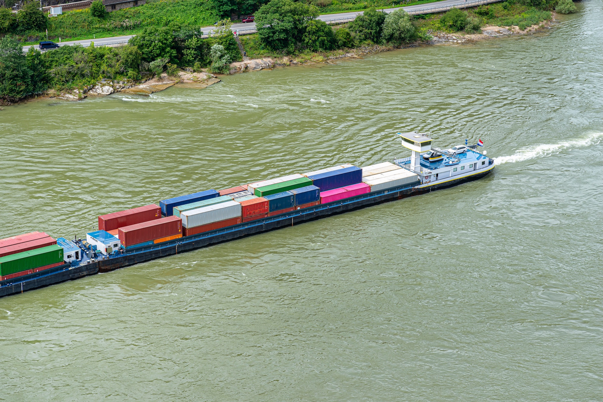 A container ship barge sailing on the river Rhine in western Germany, visible road, aerial view.