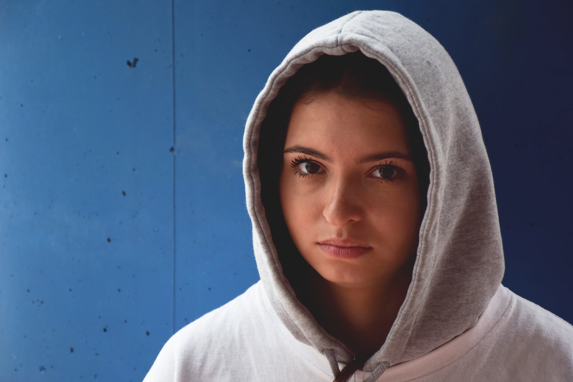 Portrait of troubled teenage girl in hoodie standing against blue wall looking at camera